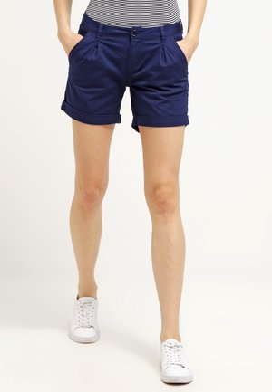 Persoon die marineblauwe, omgeslagen korte broek met zakken, een gestreept shirt en witte sneakers draagt, staand tegen een effen achtergrond.