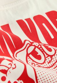 Bavlnené tričko s veľkým červeným grafickým potlačou, na ktorom sú tváre v karikatúre a výrazné písmo na krémovom pozadí; hladká textura.