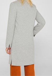 Mujer vestida con un abrigo largo gris claro con aberturas laterales y pantalones naranjas, de pie con una mano en la cadera, de espaldas a la cámara.