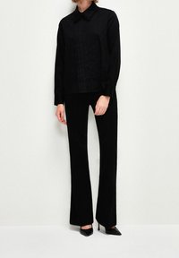 Camicia nera con colletto e pieghe verticali; abbinata a pantaloni neri a campana e tacchi a punta, il tutto realizzato in un tessuto liscio.