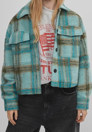 Frau trägt eine kurze türkis-braune karierte Jacke über einem grauen grafischen Sweatshirt und schwarzen Jeans, steht mit entspannten Händen.