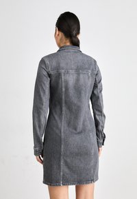 Grå denimskjortekjole med lange ermer, knappeløsninger og en tettsittende silhuett. Har en kragehals og synlige sømdetaljer.