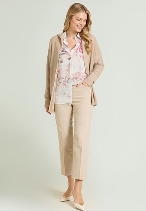 Mujer con pantalones beige, blusa floral, cárdigan topo y tacones crema con detalles dorados, de pie con una mano en el bolsillo, sonriendo y mirando hacia abajo.