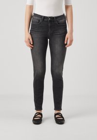 Vaqueros skinny de mezclilla negros con una cintura de altura media, bolsillos frontales y un sutil detalle de desgaste. Combinados con zapatos planos negros.