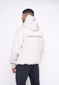 Hoodie taille oversize gris clair avec un cordon de serrage et une texture douce. Présente une embellissement textuel au dos dans des couleurs contrastantes. Associé à un pantalon sombre.