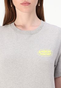 Pilkas medvilnės marškinėlis su apvaliu kaklu. Kairėje krūtinės pusėje yra neoninės geltonos spalvos "American Vintage" logotipas. Lengvas tekstūra.
