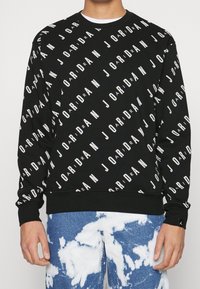 Sudadera negra con el texto "JORDAN" en blanco repetido en patrones diagonales. Puños y dobladillo acanalados, hecha de tela texturizada.