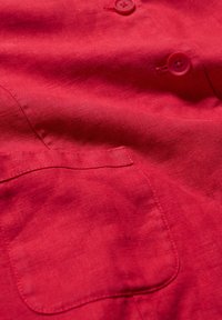 Red linen fabric featuring a smooth texture with a single chest pocket and two matching buttons. Minimalist design with clean lines.