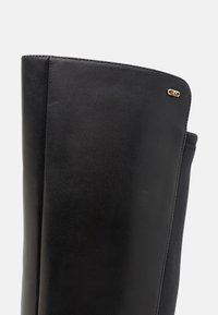 Stivale nero in pelle altezza ginocchio con una texture liscia, caratterizzato da una cucitura e un piccolo logo dorato applicato sul bordo superiore.