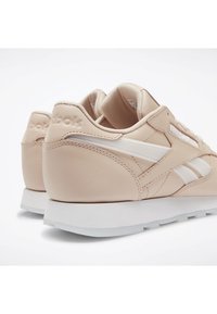 Paire de baskets beige Reebok avec des rayures blanches, des semelles blanches et des colliers rembourrés, montrée de dos et de côté sur fond blanc.