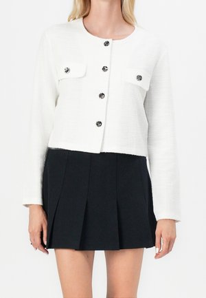 Femme portant une veste blanche texturée à boutons avec deux poches avant et une mini jupe plissée noire.