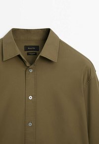 Olivgrünes Baumwollhemd mit klassischem Kragen, frontaler Knopfleiste und subtiler Textur. Verfügt über ein Markenlabel und kontrastierende Knöpfe.