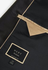 Tessuto di poliestere nero con texture a rete, caratterizzato da un bordo e un'etichetta in raso color sabbia. Include dettagli come un gancio e un'etichetta con la taglia.