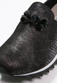 Chaussure noire texturée à enfiler avec des ornements de pierres facettées noires sur le dessus et une semelle bordée de blanc avec un dessous rainuré.