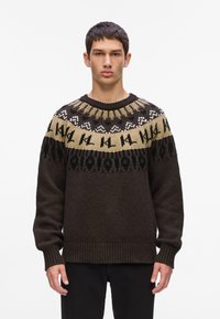 Tamno smeđi pleteni pulover s okruglim izrezom. Ima uzorkovani dio na ramenu u bež, crnoj i bijeloj boji s geometrijskim dizajnom. Rukavi su završeni rebrastim detaljima.