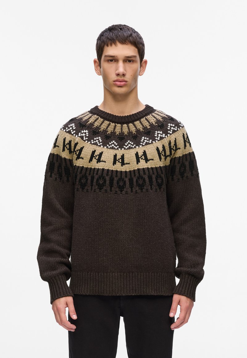 Tamno smeđi pleteni pulover s okruglim izrezom. Ima uzorkovani dio na ramenu u bež, crnoj i bijeloj boji s geometrijskim dizajnom. Rukavi su završeni rebrastim detaljima.