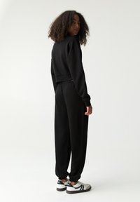 Hanorac negru scurt cu mâneci lungi și talie elastică, asortat cu pantaloni jogger negri. Modelul poartă adidași albi cu accente negre.