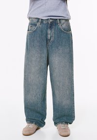Persona che indossa jeans azzurri larghi e sneakers beige, in piedi contro uno sfondo bianco dalla vita in giù.