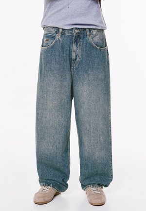 Person, der locker sitzende blaue Jeans und beige Sneakers trägt, steht mit dem Unterkörper vor einem weißen Hintergrund.