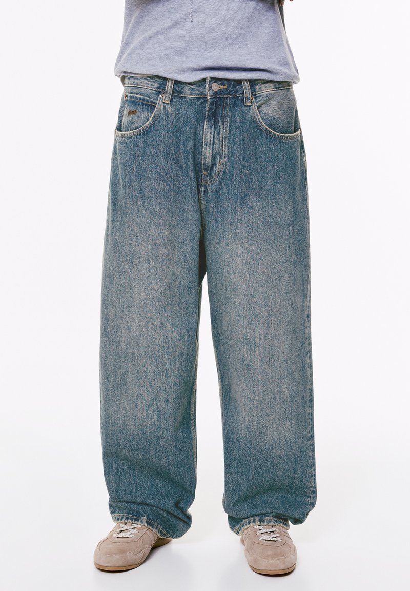 Persona che indossa jeans azzurri larghi e sneakers beige, in piedi contro uno sfondo bianco dalla vita in giù.