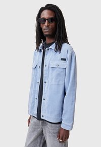 Chaqueta de pana azul claro con botones a presión, dos bolsillos en el pecho y un parche negro con logo. Llevada sobre una camiseta negra y unos jeans grises.