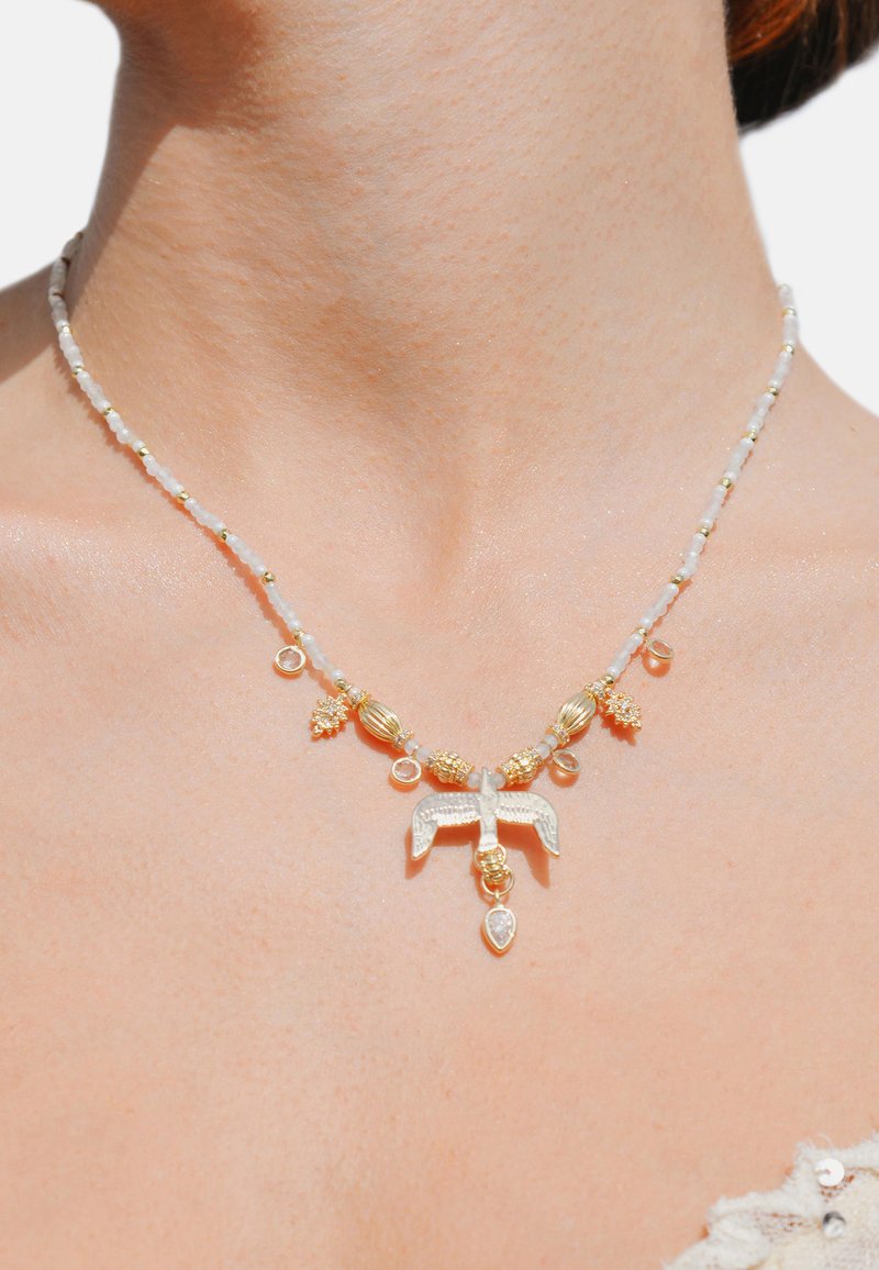 Collier mettant en avant un pendentif central en argent en forme d'oiseau, des perles dorées et blanches, ainsi que de petits accents décoratifs. Les perles sont translucides et présentent une texture lisse.