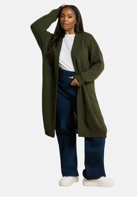 Lång olivgrön kofta med strukturerad stickning,  öppen framsida och sänkta axlar, parat med mörkblå vida jeans och vita sneakers.