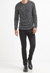 Muž nosí černé a bílé skvrnité triko s dlouhým rukávem, černé slim kalhoty a černé kožené společenské boty, které kráčí vpřed.