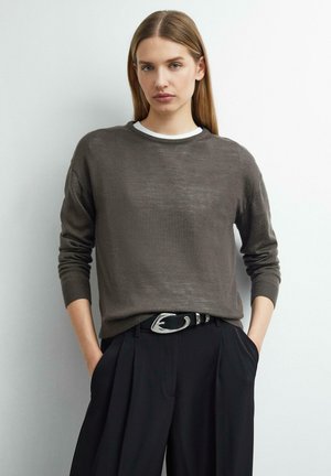 Jeune femme aux cheveux raides portant un pull marron sur une chemise blanche et un pantalon plissé noir avec une ceinture boucle argent.