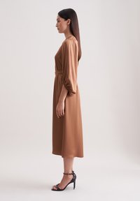 Braunes knielanges Kleid mit tailliertem Schnitt und puffärmel, aus glattem Stoff. Dazu werden schwarze High-Heel-Sandalen getragen.