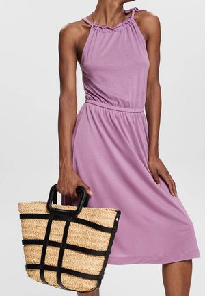 Woman wearing sleeveless lilac dress with gathered neckline, holding woven straw tote bag with black trim and handles.