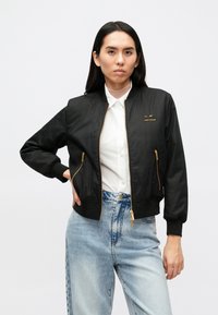 Μαύρο μπουφάν bomber με χρυσά φερμουάρ, ριμπ μανσέτες και λογότυπο στο στήθος. Φορεμένο πάνω από λευκό πουκάμισο και τζιν ανοιχτού χρώματος.