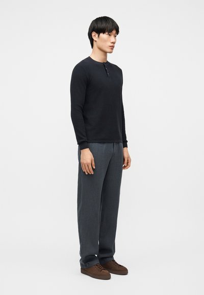 Άνδρας που στέκεται με μαύρο πουκάμισο μακρυμάνικο, γκρι παντελόνι και καφέ παπούτσια μπροστά σε απλό φωτεινό φόντο, κοιτάζοντας προς τα αριστερά.