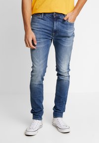 Džínové kalhoty v bleděmodré barvě se slim fit, pěti kapsami a zapínáním na knoflík. Nosené s bílými teniskami a žlutou košilí.