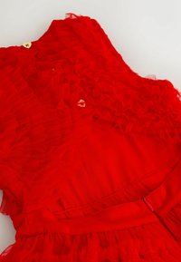 Červené šaty so vrstvenými volánmi, jemnou textúrou a priliehavým pásom. Výrazné detaily zahŕňajú zlaté gombíky v tvare srdca a skrytý zips.