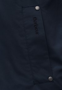 Ύφασμα μπουφάν σε ναυτικό μπλε, με ραμμένο λογότυπο "Barbour" και ασημένιο κουμπί με σφραγισμένες λεπτομέρειες. Λείο υφή και μινιμαλιστικός σχεδιασμός.