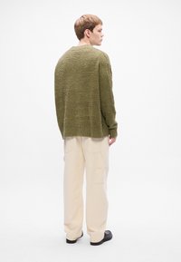 Maglione verde oliva testurizzato con una vestibilità ampia, orlo a costine e spalle abbassate, abbinato a pantaloni a gamba larga color crema e scarpe nere.