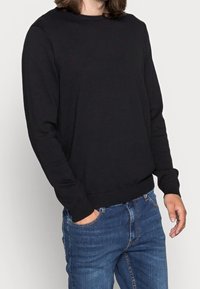 Pull en tricot noir à manches longues, col rond et bords-côtes, associé à un jean en denim bleu. Texture lisse, coupe classique.