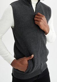 Gilet grigio a maglia con zip frontale, collo alto e texture a coste. Presenta tasche laterali ed è indossato sopra una camicia bianca a maniche lunghe.