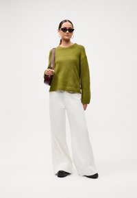 Ženska oblečena v olivno zeleni pulover, široke bele hlače, črne čevlje, sončna očala v obliki mačjih oči, uhane s krogom in nosi burgundsko torbico čez ramo.