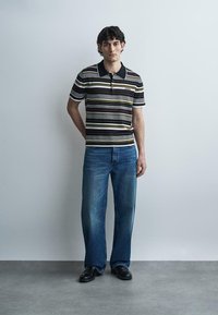 Νέος άνδρας που στέκεται σε έναν απλό τοίχο φορώντας ριγέ πόλο, μπλε τζιν και μαύρα παπούτσια.