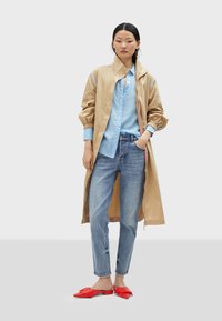 Beige, lightweight long jacket with elastic cuffs, layered over a light blue button-up shirt with polka dots, paired with blue jeans and red flat shoes.