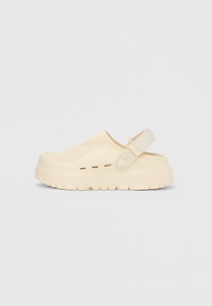 Chaussure beige à enfiler en matériau souple, avec un bout arrondi, une tige texturée et une bride arrière pour un maintien sécurisé. Semelle épaisse rainurée.