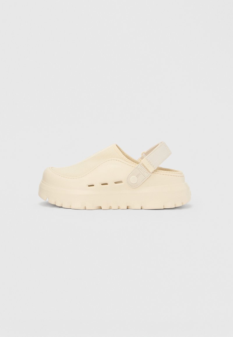 Chaussure beige à enfiler en matériau souple, avec un bout arrondi, une tige texturée et une bride arrière pour un maintien sécurisé. Semelle épaisse rainurée.