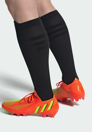 Bright orange soccer cleats feature a textured upper with green stripes and three stripes logo. Black knee-high socks have ribbed design.