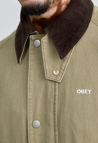 Khaki jack met een bruine corduroy kraag, voorzien van een metalen drukknop en het geborduurde "OBEY" logo in het wit op de stof.