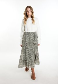 Witte, gestructureerde trui met lange mouwen, gecombineerd met een gelaagde rok met bloemenpatroon in donkergroen en crème. Bruine laarzen tot de knie.