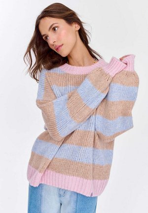 Kobieta w luźnym, paskowanym swetrze w odcieniach beżu, niebieskiego i różowego, w jasnoniebieskich dżinsach, pozująca z rękami uniesionymi blisko twarzy.
