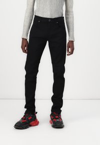Personne portant un pantalon slim noir, un haut gris côtelé à manches longues et des baskets noires avec des touches rouges, debout devant un fond blanc uni.