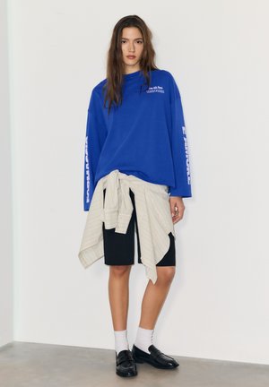 Blauwe trui met lange mouwen en witte tekst, zwarte shorts, licht gestreepte wikkelrok vastgebonden in de taille, en zwarte loafers met witte sokken.